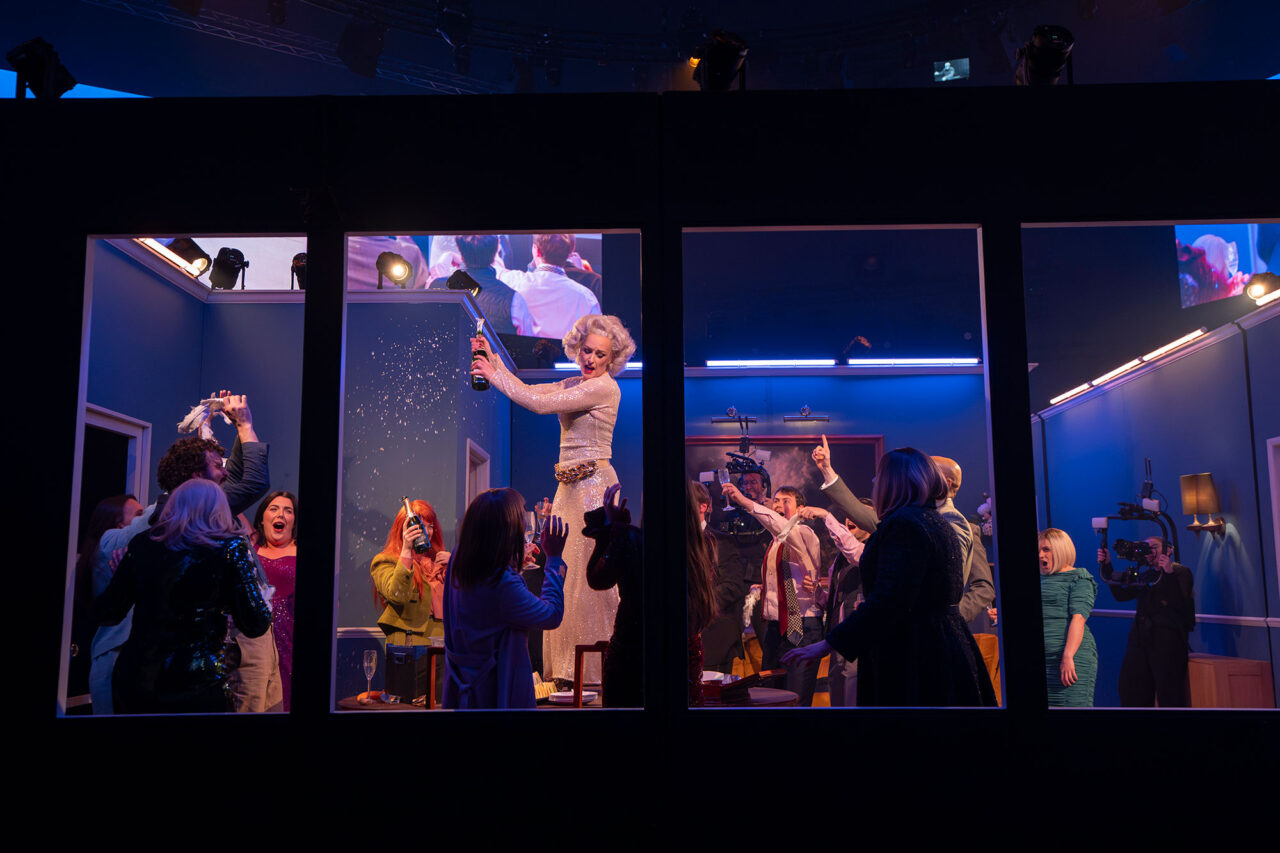 A lively party scene viewed through large windows shows a woman in a sparkling dress standing on a table, opening a bottle and spraying champagne, surrounded by cheering guests taking photos and celebrating in a blue-lit room.