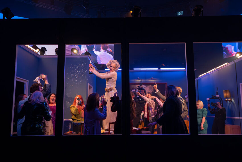 A lively party scene viewed through large windows shows a woman in a sparkling dress standing on a table, opening a bottle and spraying champagne, surrounded by cheering guests taking photos and celebrating in a blue-lit room.