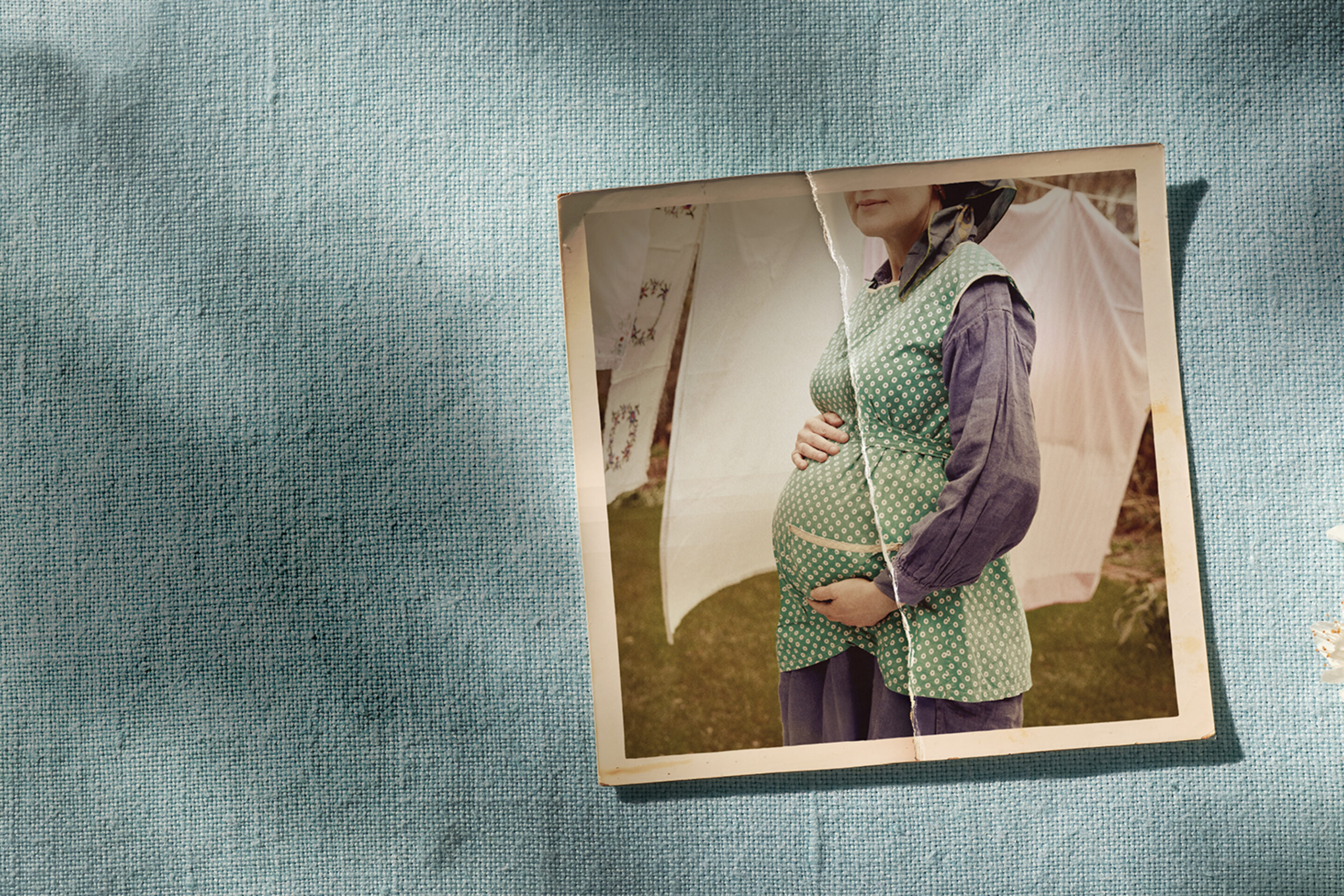 A vintage photo of a pregnant woman wearing a green polka dot waistcoat and blue dress, holding her belly, is placed on textured blue fabric next to blossoming white flowers. The woman's face is partially out of frame.