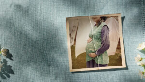 A vintage photo of a pregnant woman wearing a green polka dot waistcoat and blue dress, holding her belly, is placed on textured blue fabric next to blossoming white flowers. The woman's face is partially out of frame.