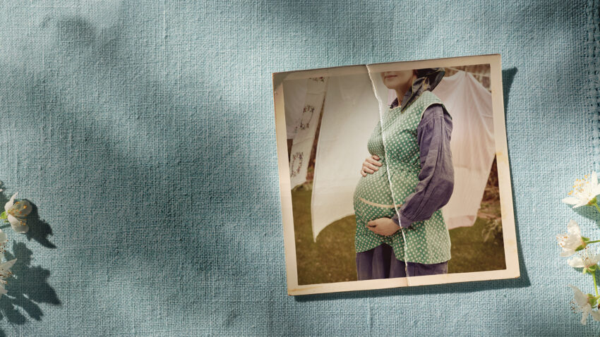 A vintage photo of a pregnant woman wearing a green polka dot waistcoat and blue dress, holding her belly, is placed on textured blue fabric next to blossoming white flowers. The woman's face is partially out of frame.