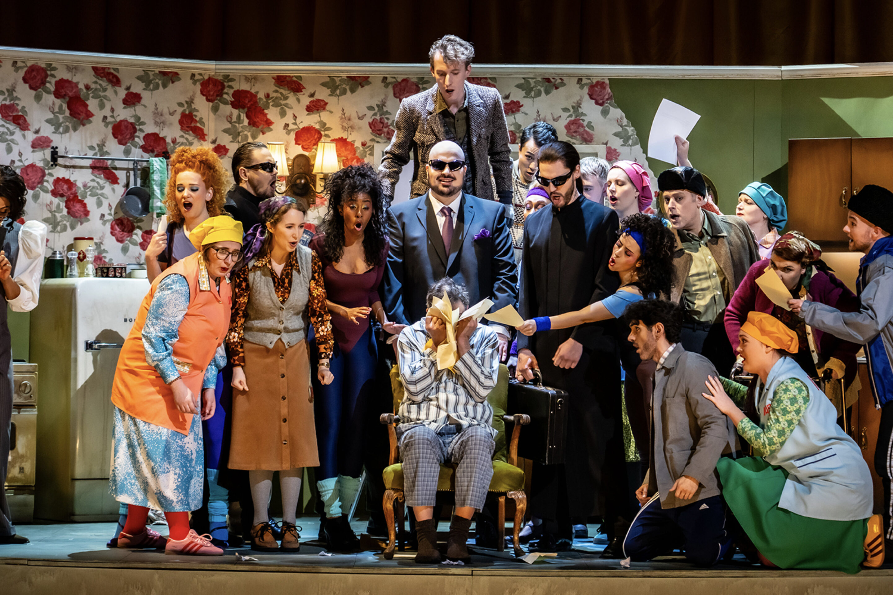 A colourful stage scene shows a group of actors in vibrant costumes gathered around a person sitting in a chair, covering their face, as others gesture and hold papers in a lively, comedy atmosphere.