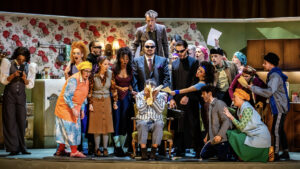 A colourful stage scene shows a group of actors in vibrant costumes gathered around a person sitting in a chair, covering their face, as others gesture and hold papers in a lively, comedy atmosphere.