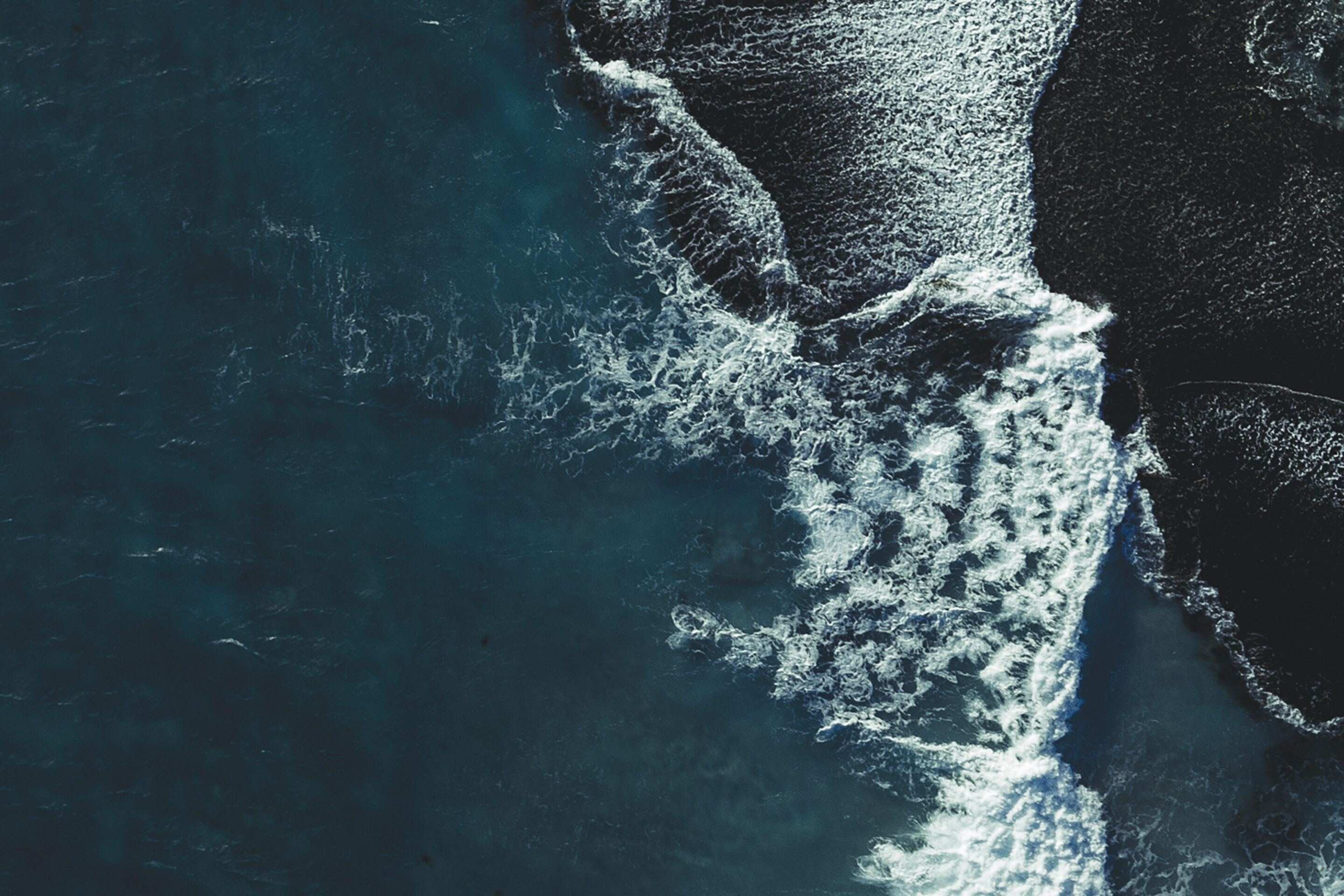 Aerial view of ocean waves crashing onto dark, textured sand, creating striking white foam patterns and deep contrasts along the shore.