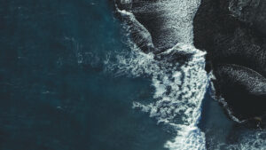 Aerial view of ocean waves crashing onto dark, textured sand, creating striking white foam patterns and deep contrasts along the shore.