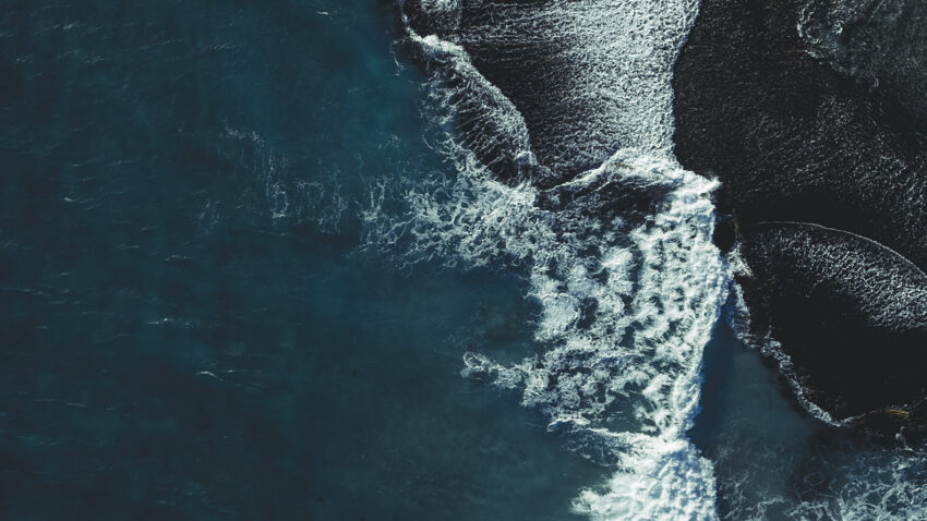 Aerial view of ocean waves crashing onto dark, textured sand, creating striking white foam patterns and deep contrasts along the shore.