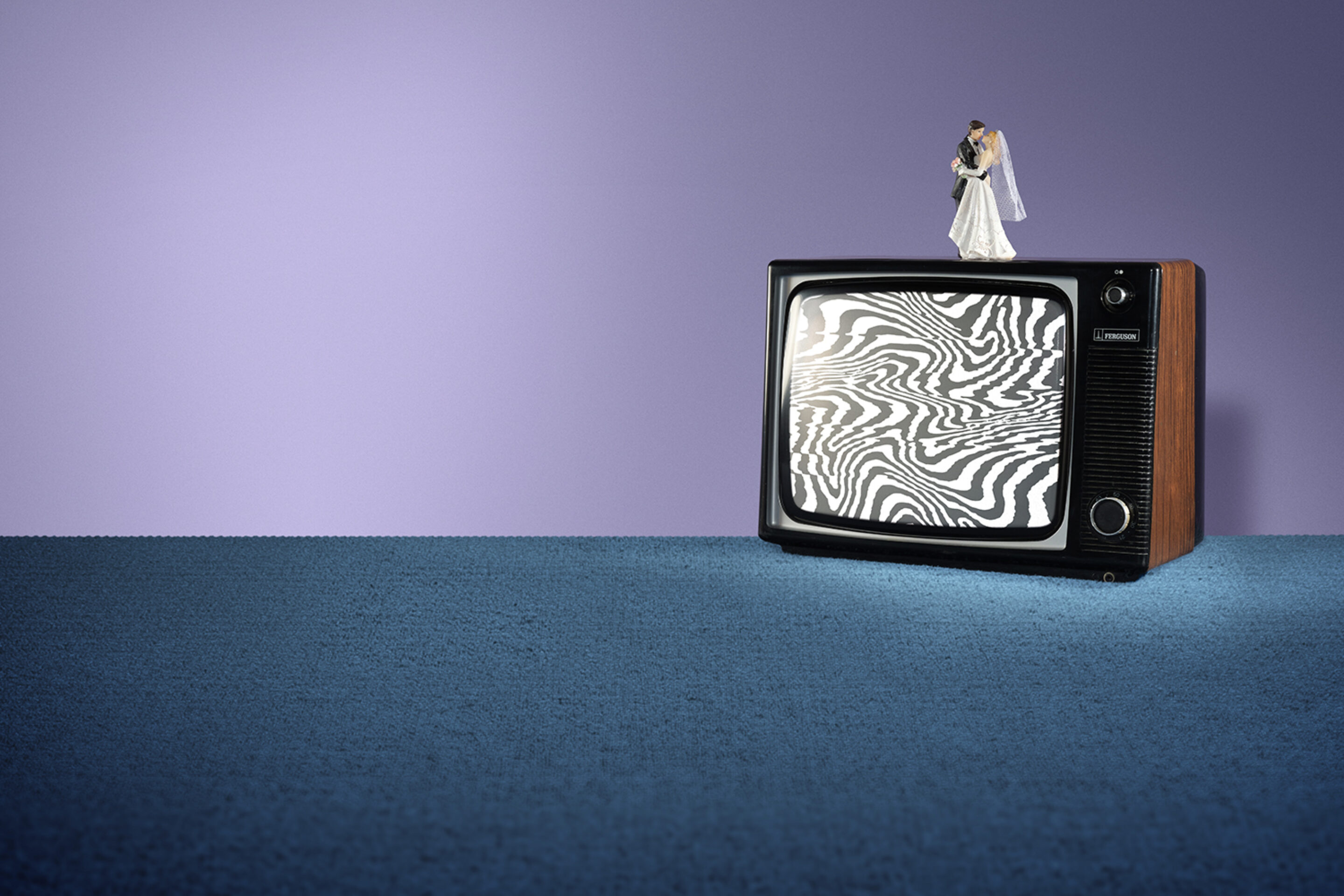 A vintage TV with static, wavy black-and-white lines on the screen sits on blue carpet. On top of the TV is a bride and groom wedding cake topper, set against a purple background.